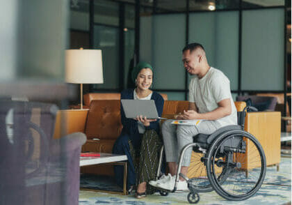 A man in a wheelchair and a woman sitting next to him showing something on a laptop