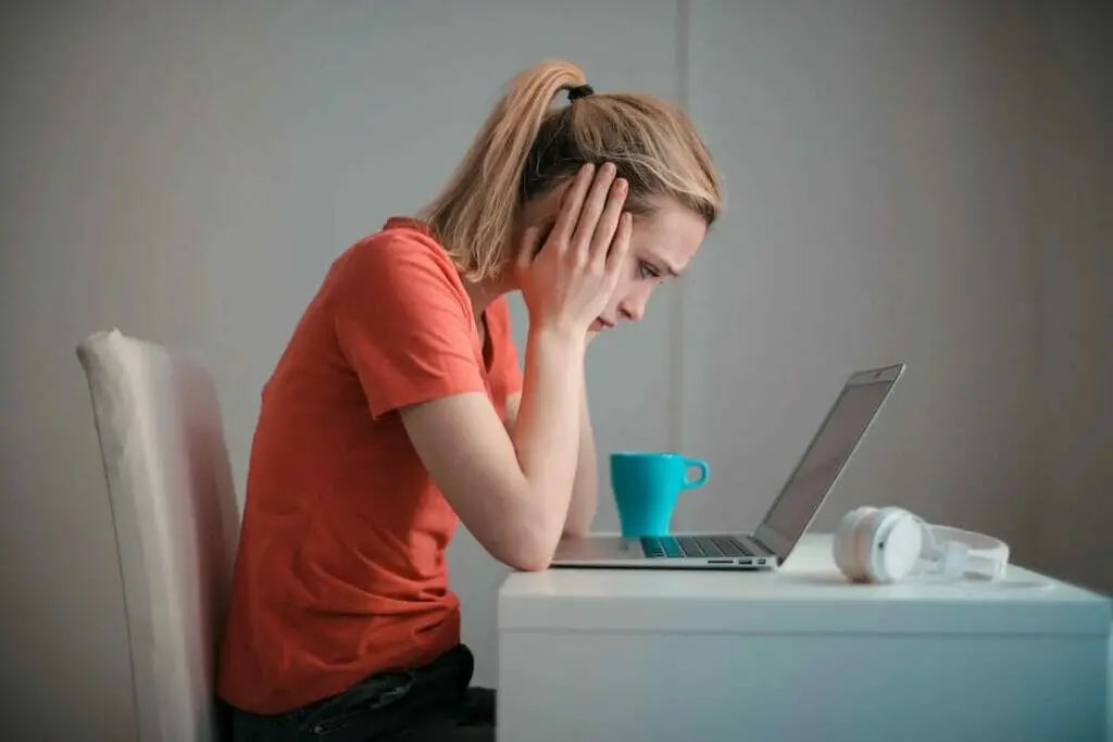 A woman holding her face is sitting at a desk with a laptop and a cup of coffee