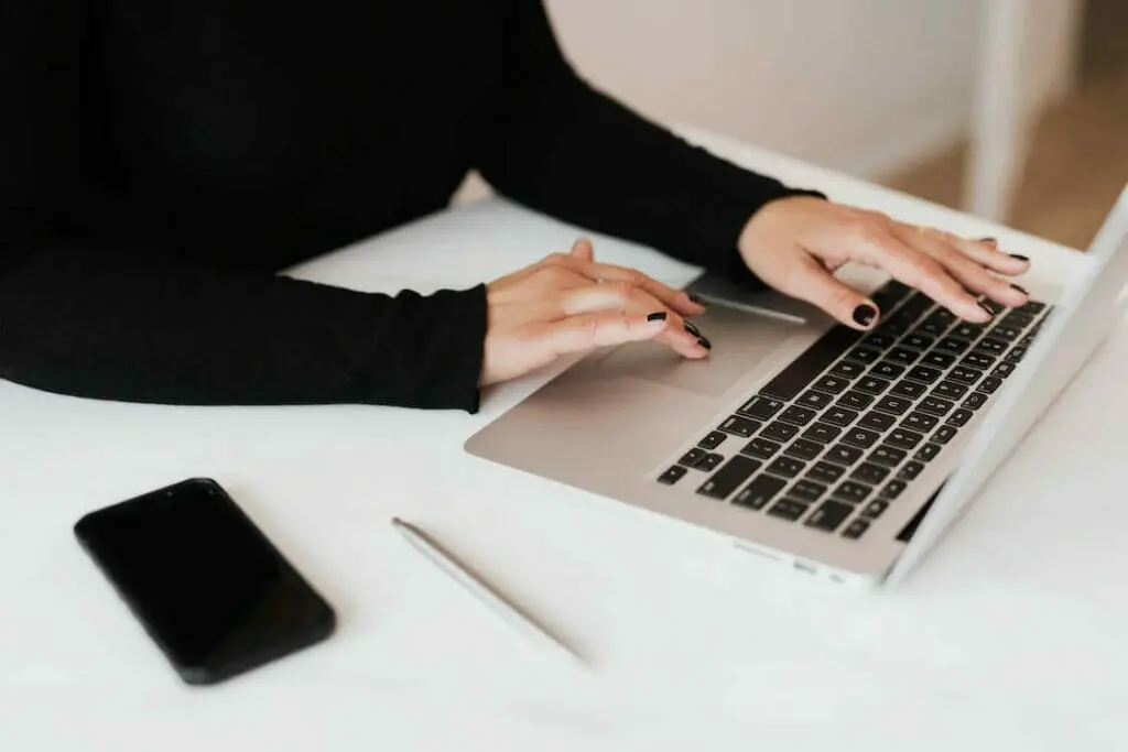 A woman typing on a laptop with a cell phone