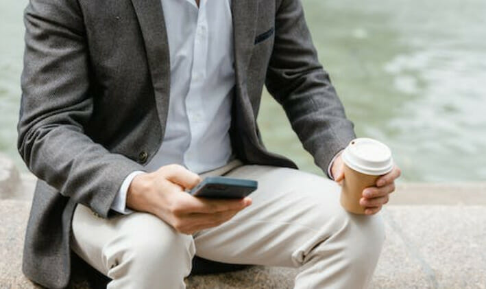 A man sitting near the body of water while holding a cup of coffee and a cell phone