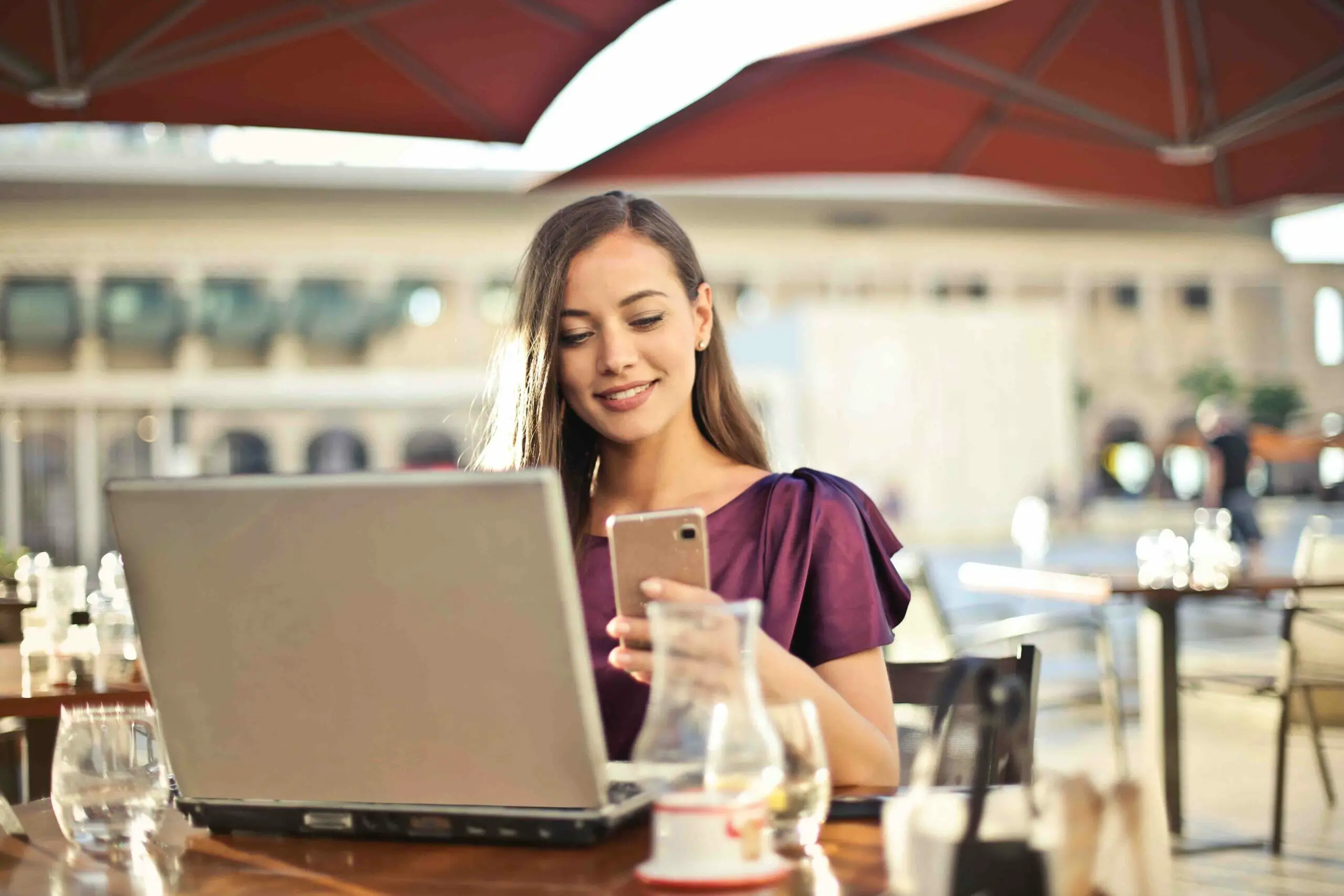A woman at the coffee shop smiling while holding her phone and an open laptop on the table