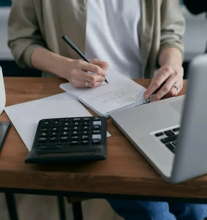 A person sitting on the desk writing on the notebook in front of a laptop, calculator, and blank paper