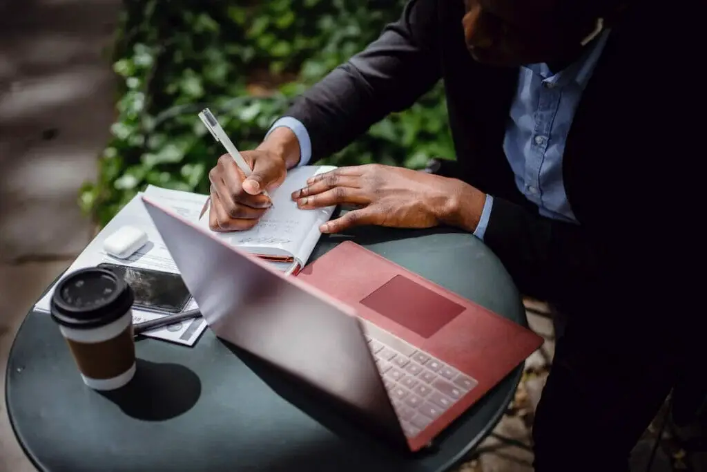 A man sitting at the outdoors writing on his notebook while checking on his laptop