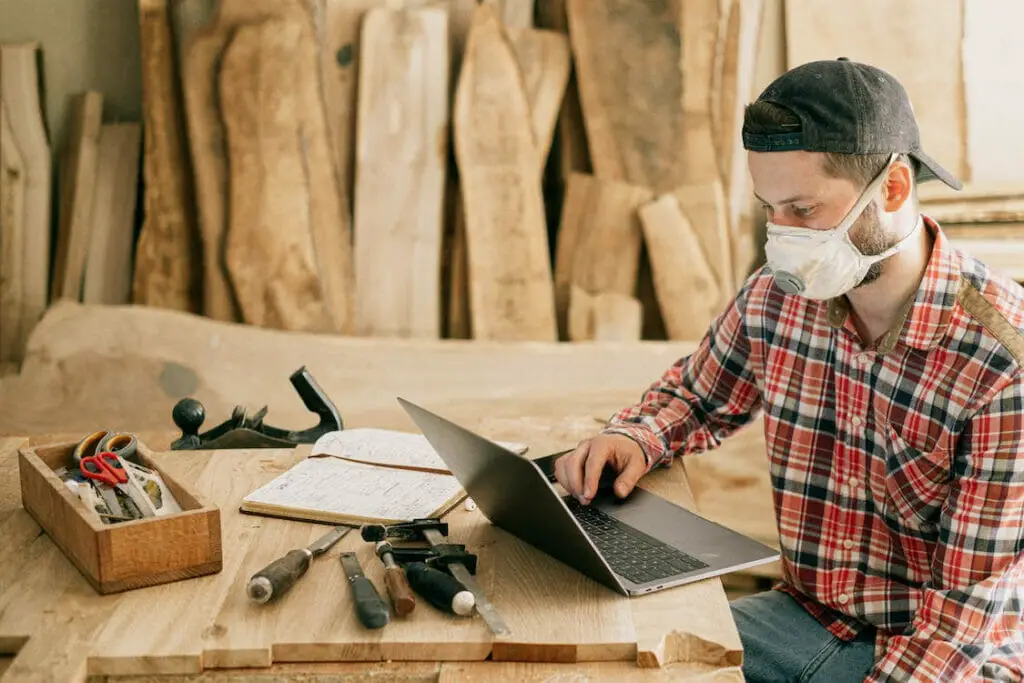 A man in a mask sitting at a lumber house using his phone at the table while surrounded by tools