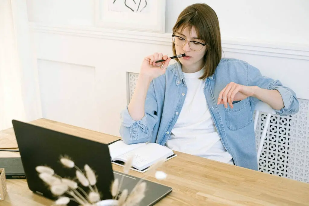 A woman sitting on the table facing her laptop with her pen on the mouth