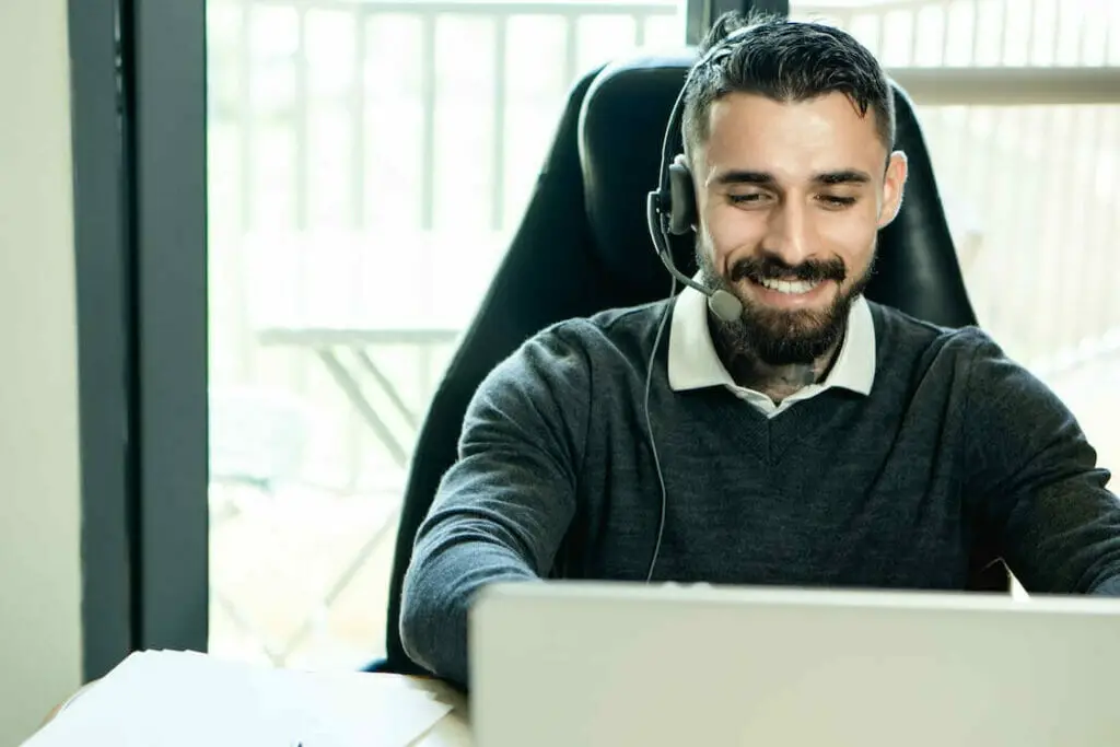 A man wearing a headset is smiling while working on a laptop