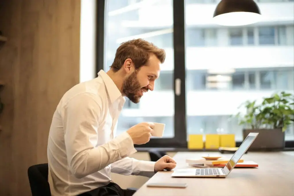 A man in his office desk holding a cup of coffee while working on his laptop